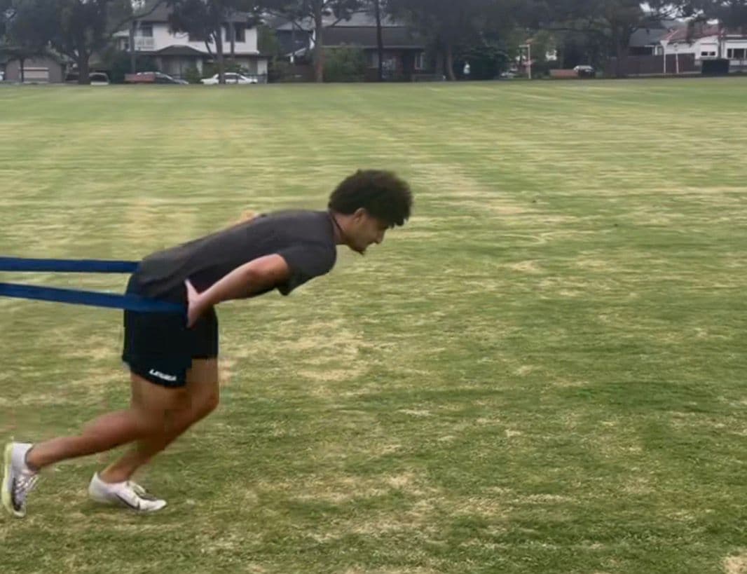 Adam in resistance-band-loaded drive-phase position — forward lean, full triple extension, driving horizontally into the ground